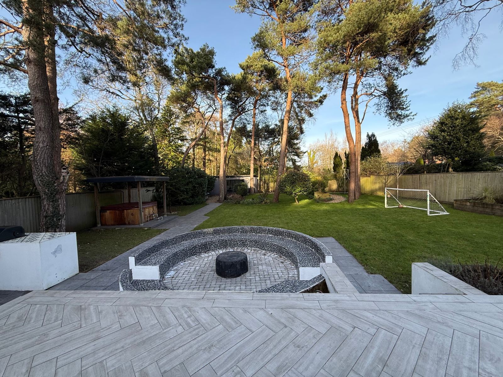 The garden at Kiwi Pines: a sunken fire pit with curved seating, lawn with a football goal, and the covered hot tub under tall pine trees